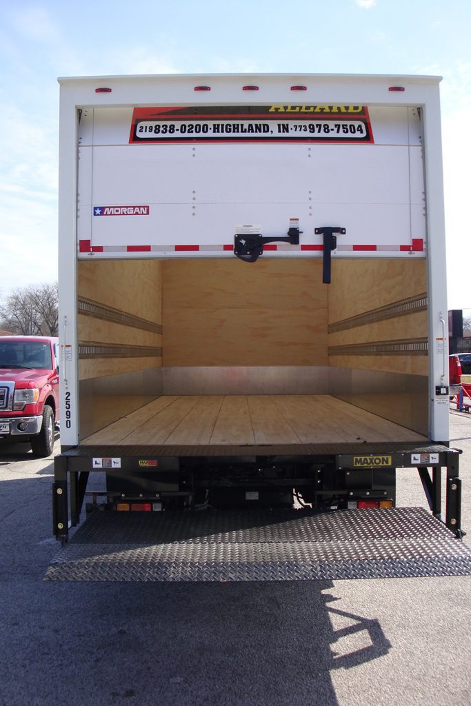 Who We Serve Open empty box truck with wooden interior and liftgate, parked on asphalt with other vehicles visible in the background.