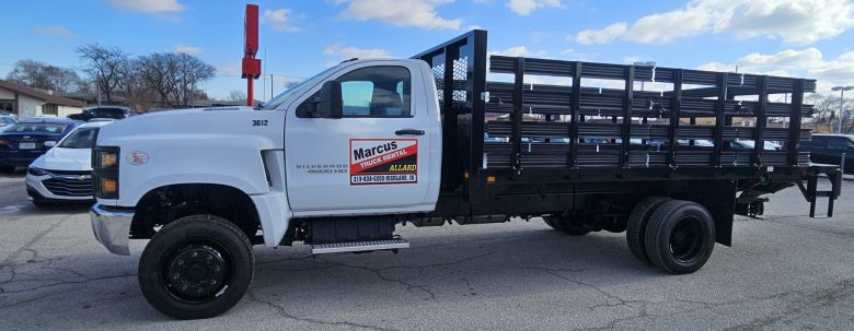 White flatbed truck with a metal rack, parked in a lot under a blue sky with clouds. Advertisements for "Hammond 8 Passenger Van Rental” and a red contact number are visible on the driver's door.