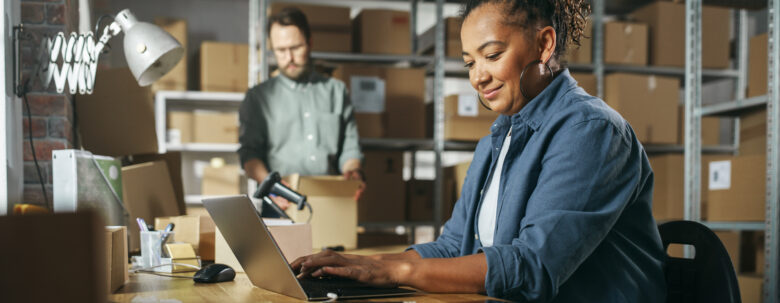 Woman uses a laptop at a desk in a warehouse office, while a man packs boxes in the background—possibly preparing an order for truck rental Merrillville. Shelves with cardboard boxes line the room.